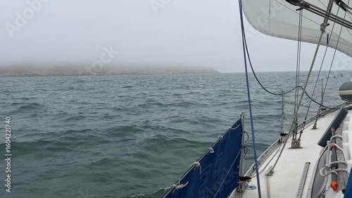A sailing boat gently going downwind in light airs approaching a headland in fog. A sailing yacht in restricted visibility, calm seas, under autopilot.