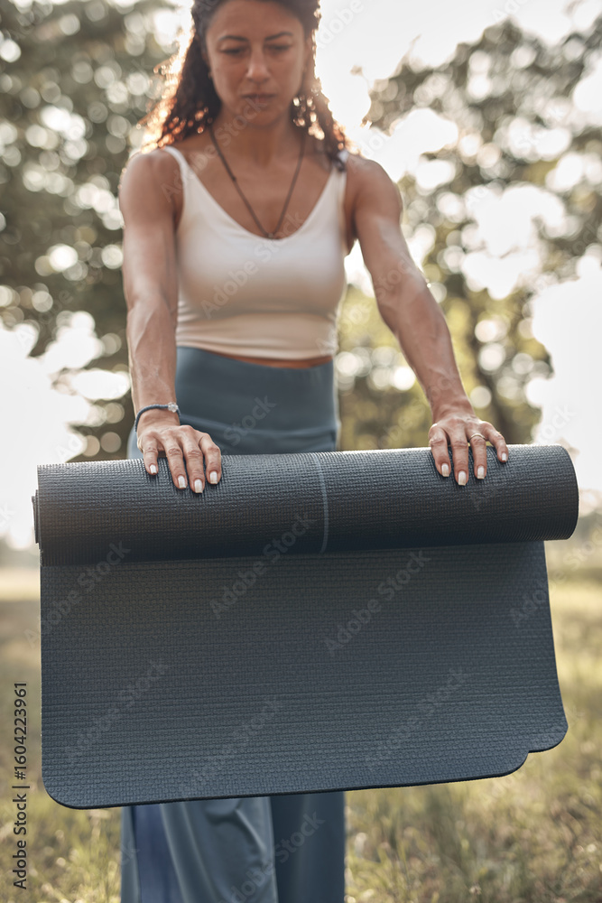 Fototapeta premium Woman folding and unfolding yoga mat in the park during sunset sunrise time.