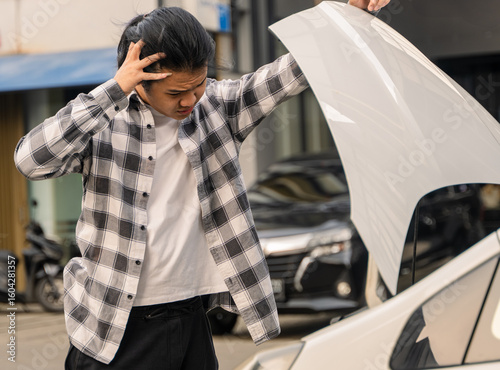 Asian man looking frustrated while checking under the hood of a white car, standing on a city street with his hand on his head, possibly dealing with car trouble or a breakdown