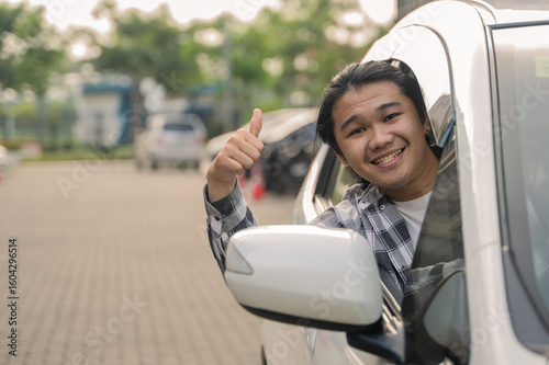 Asian man in a checkered shirt smiles and gives a thumbs up while leaning out of a car window in an outdoor parking area