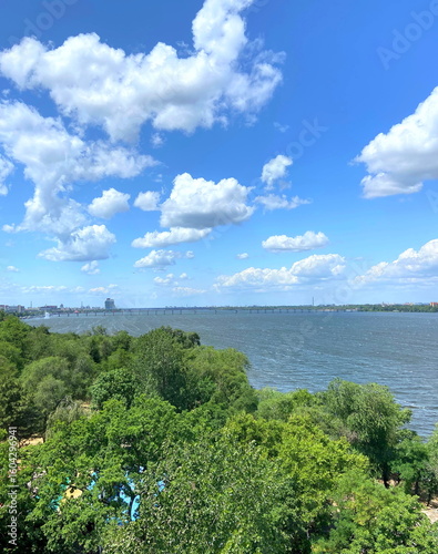 View from the Ferris Wheel of a dense coastal forest strip along the wide Dnipro riverbed against a blue sky covered with illuminated clouds.