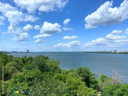 Landscape from the Ferris Wheel of a long automobile bridge across the Dnipro against a flock of clouds in a blue sky illuminated by the sun's rays.