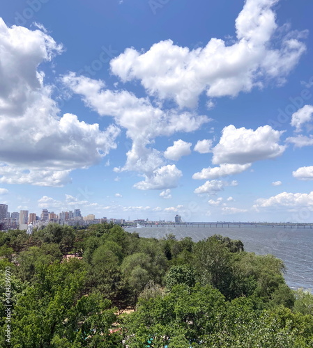 View from the Ferris Wheel of a dense coastal forest strip along the wide Dnipro riverbed against a blue sky covered with illuminated clouds.