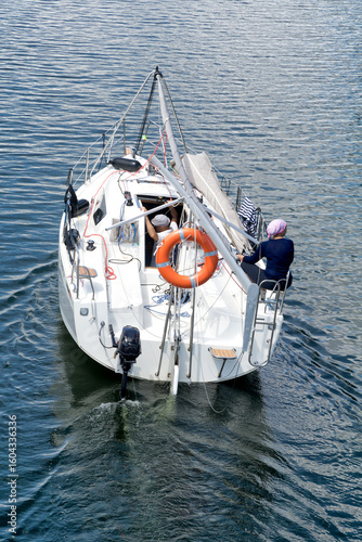A sailboat maneuvers powered by the engine without sails, with the mast lowered, rear view. Two sailors on board. Leasure, travelling, vacations.