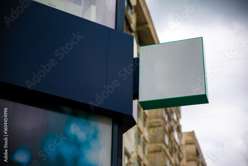 Empty rectangular store sign mounted on modern building with city apartments in background and cloudy sky above.