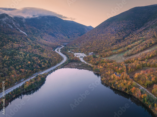 Aerial view of the winding road mirroring the serene lake's edge, nestled between mountains ablaze with autumn colors, Franconia Notch State Park, Vermont, United States.