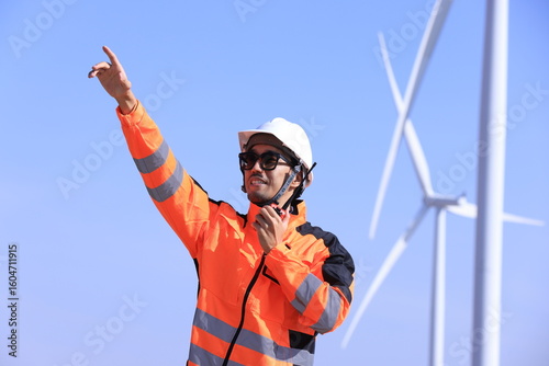 Engineer or architect with helmet working with laptop for inspection details on construction site with wind turbine farm sunset background. Asian foreman working in renewable energy business. 