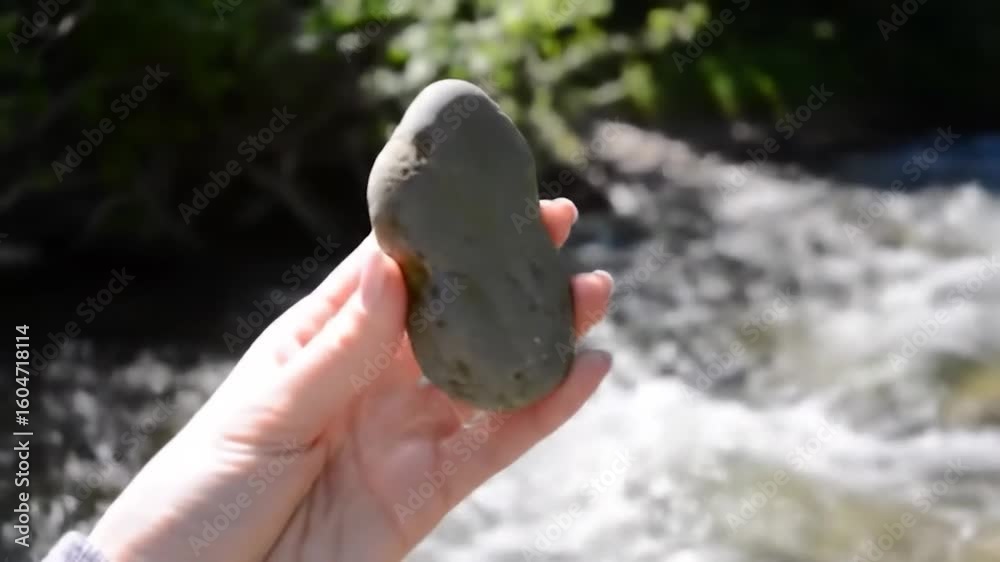 Hand holds a smooth, gray river stone against a blurry background of flowing water and greenery