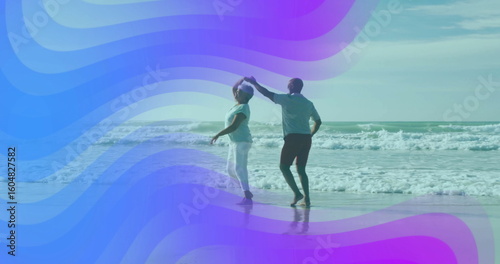 Dancing mature couple twirling barefoot at sandy beach, with rolling ocean waves and shallow water
