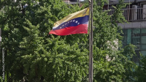Slow motion of Venezuelan flag moving in wind surrounded by green trees and sunlight in international district