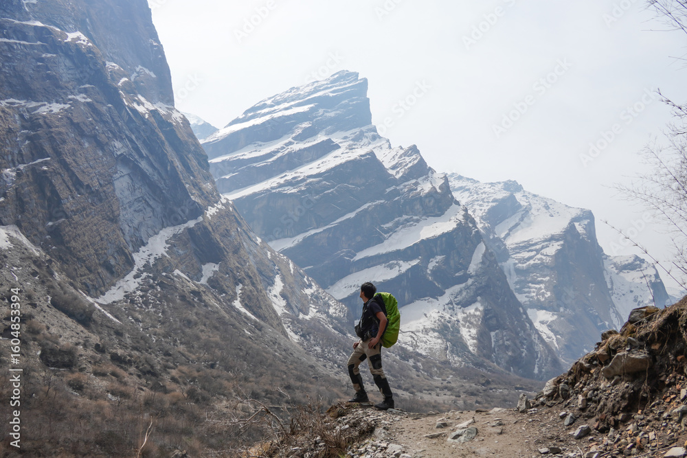 Fototapeta premium Young asian male hiker carrying mountain backpack and using trekking pole took photos after successfully hiking to Annapurna Base Camp, with the snow-capped Himalayan peaks visible in the morning