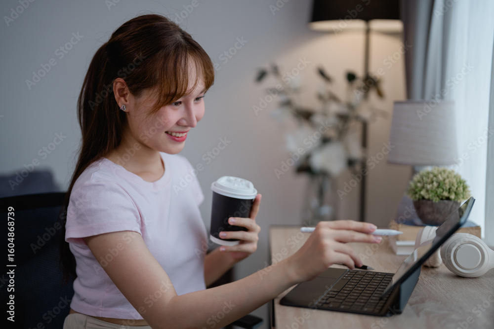 Smiling young Asian freelancer woman working remotely from home, savoring a cup of coffee while using her tablet with a keyboard and stylus pen in a cozy, well-lit living room Smiling young Asian freelancer woman working remotely from home, savoring a cup of coffee while using her tablet with a keyboard and stylus pen in a cozy, well-lit living room