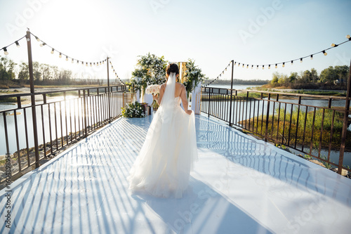 A brunette bride in a white dress holds a bouquet and walks to the wedding arch, among many white chairs. Wedding ceremony in nature. Celebration