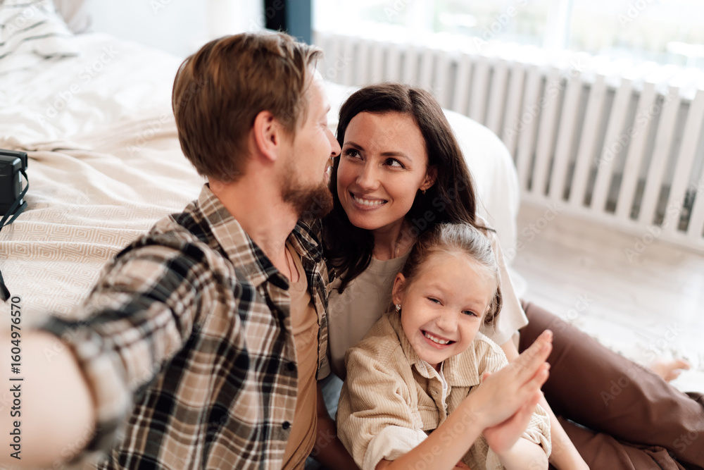 Obraz premium Happy family smiles while father takes selfie, daughter clapping hands joyfully, mother looking lovingly, tender moment filled with fun, warmth, and connection.