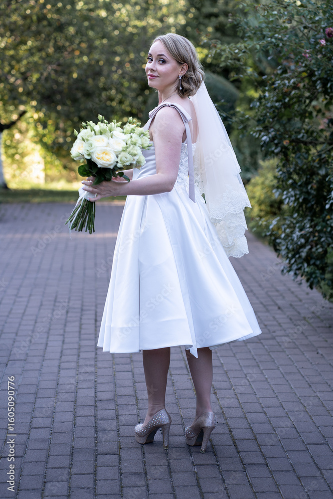 Stylish bride in a tea-length white wedding dress and lace veil, holding a bouquet of ivory and blush roses with green buds. Full-length outdoor portrait showcasing elegant high heels, soft natural Stylish bride in a tea-length white wedding dress and lace veil, holding a bouquet of ivory and blush roses with green buds. Full-length outdoor portrait showcasing elegant high heels, soft natural