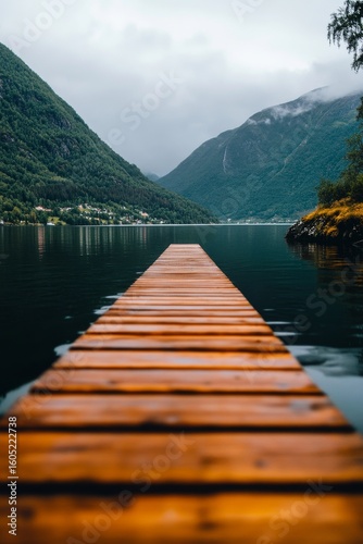 Wooden Dock Extending Over Calm Lake Surrounded by Mountains Under Cloudy Sky.