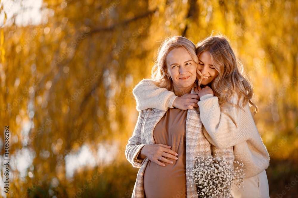 Fototapeta premium Adult daughter hugs her pregnant mom while standing in autumn park
