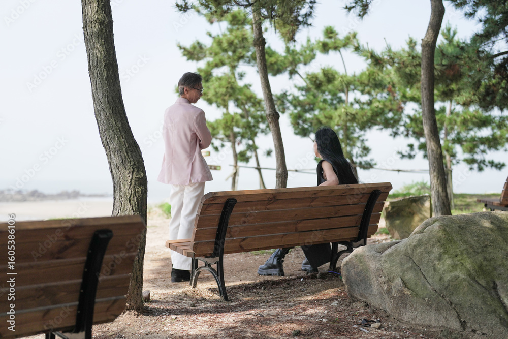 Fototapeta premium In June 2025, a Korean man and woman in their late fifties sit on a bench by the sea, chatting and smiling as they enjoy the breeze, ocean view, and calm surroundings in Incheon Metropolitan City.
