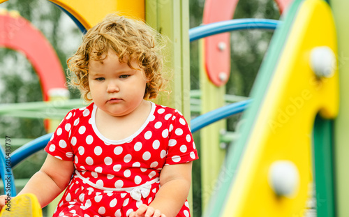 A young child with curly hair sits on a colorful playground slide wearing a red dress with white polka dots The playground is bright and cheerful contrasting with the childs somber expression