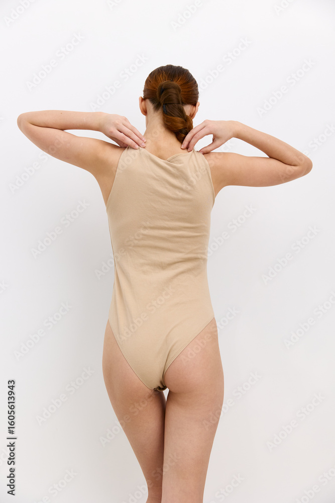 Obraz premium Woman in beige bodysuit with tied hair posing with hands near neck against plain white background, showcasing minimalist style and natural light.
