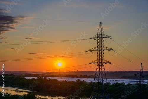 High-voltage power lines and electricity pylons at sunset over river and forest landscape, vivid orange sky, energy transmission infrastructure, industrial silhouettes, peaceful evening view