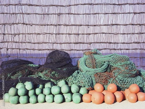 Fishing nets and buoys stacked against a woven reed fence in bright sunlight