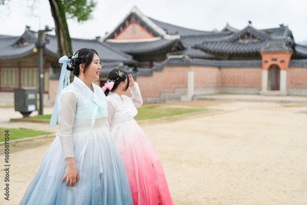 Fototapeta premium In Seoul in June 2025, two Korean women in their twenties wear pink and sky-blue hanbok as they laugh and chat in front of the ornate doors of Gyeongbokgung Palace, their joy echoing so softly.