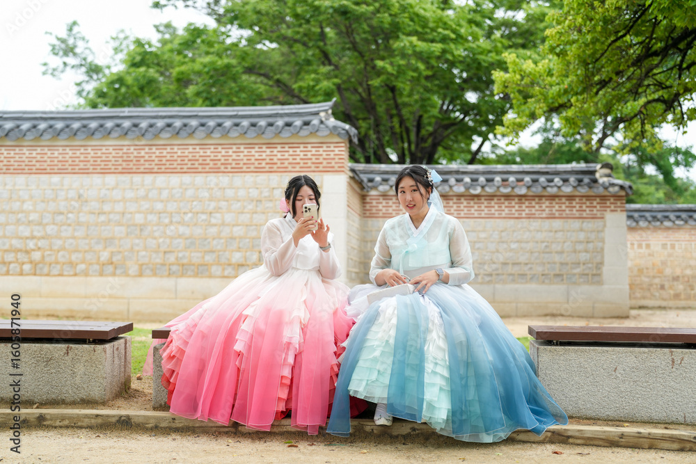 Fototapeta premium In Seoul in June 2025, two Korean women in their twenties, dressed in charming pink and sky-blue hanbok, sit in front of the traditional facade of Gyeongbokgung Palace, sharing a peaceful moment.