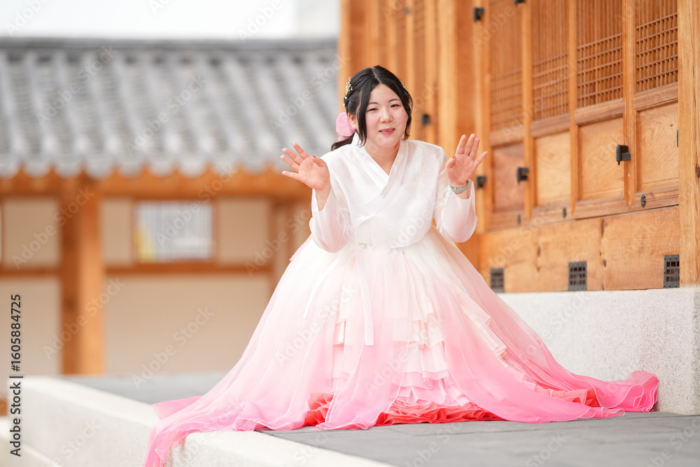 Fototapeta premium A Korean woman in her twenties wearing a charming pink hanbok sits in front of the traditional facade of Gyeongbokgung Palace, her calm demeanor reflecting timeless beauty, in Seoul in June 2025.