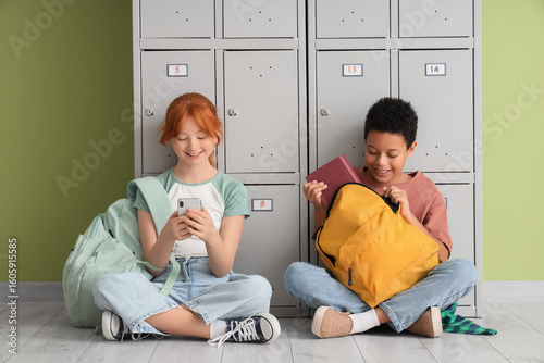 Teenage students with backpacks and mobile phone sitting near locker at school