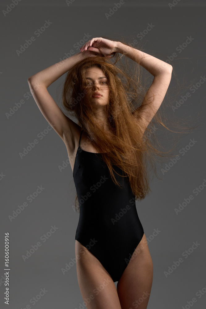 Fototapeta premium slender woman with long red hair posing confidently in black bodysuit against gray background, natural light highlighting her skin and hair texture