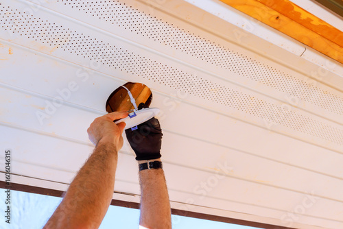 Photography Homeowner applies lighting fixture in ceiling in soffit under eaves of house in suburb