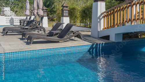 Wicker deck chairs and tables on the outdoor terrace by the swimming pool. The blue tiled bottom is visible through the clear water. Arched pedestrian bridge. Tropical vegetation. Philippines.