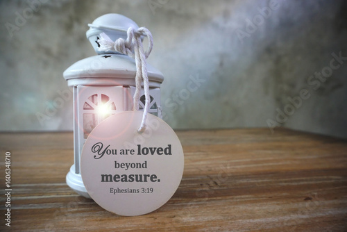 Bible verse quote - You are loved beyond measure. Ephesians 9:13 on a white circle label paper hanging with rope on classic light of lantern on the wooden table. Religious text. Christianity concept.