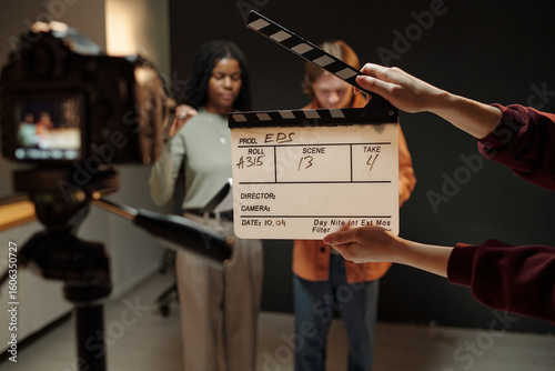 Young Black woman and young Caucasian man standing in front of camera during acting audition, holding script while crew member holding clapperboard in foreground, studio background visible