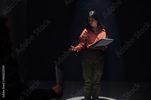 Young adult Hispanic woman standing on stage holding script, performing monologue during acting audition, gesturing with hand while looking at casting director in dark theater setting