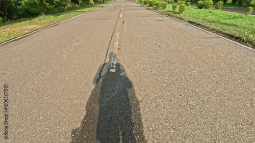 point of view of man shadow running or jogging with green trees on asphalt road in the morning