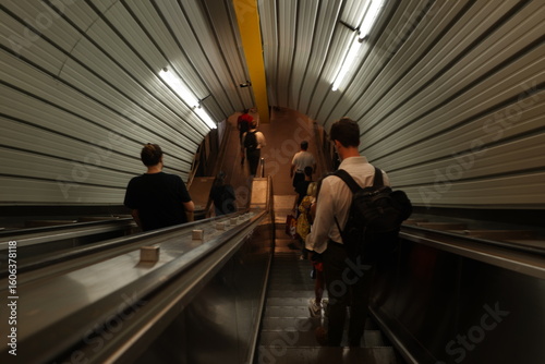 Travelers on escalators descend into a busy transit station in the Eternal City. The metallic, ribbed tunnel creates a contemporary backdrop for the moving public, highlighting the pulse of city exist