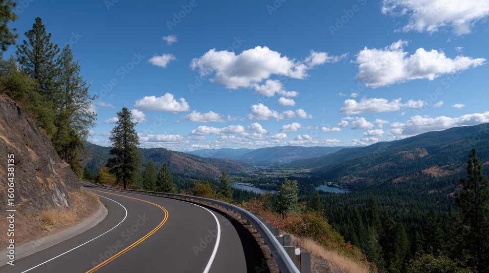 Fototapeta premium Scenic Highway Curving Through Mountain Landscape Under Bright Blue Sky with Fluffy White Clouds