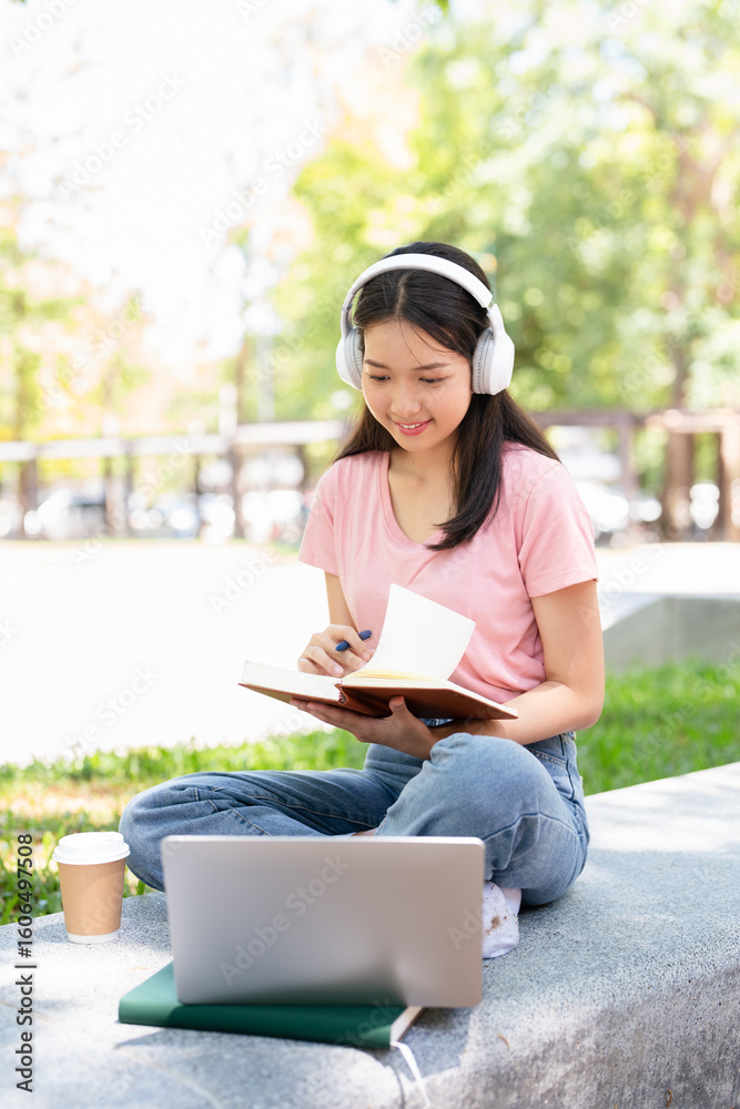 Obraz premium Smiling young asian woman in wireless headphones sitting outdoor working on laptop computer and writing or taking notes in paper notebook, holding pen in hand.