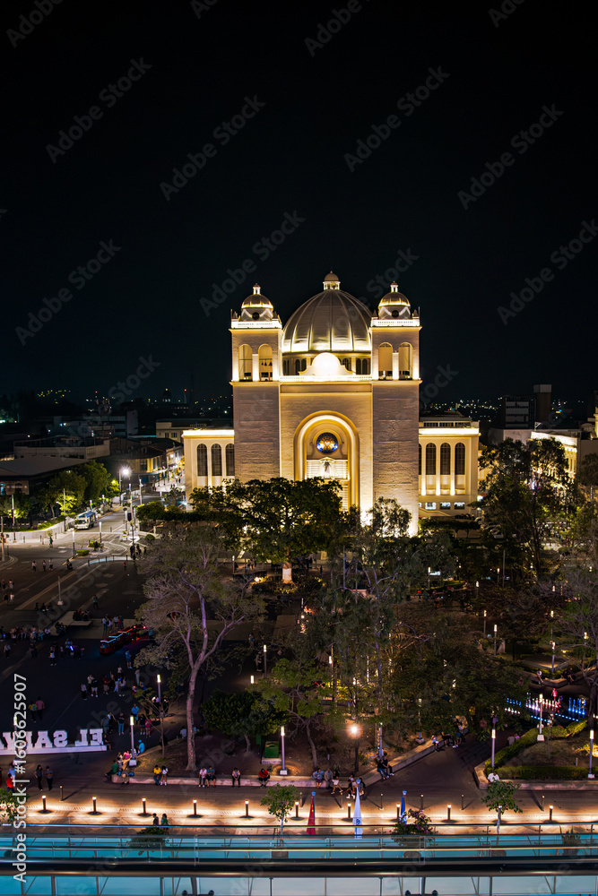 Fototapeta premium A beautifully lit cathedral or historic building stands tall at night in the heart of the city, surrounded by trees and a lively public square. Captured from above with a vibrant urban ambiance.