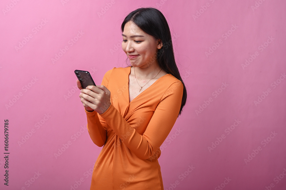 Asian woman with long black hair wearing a fitted orange dress stands indoors against pink background, holding smartphone with both hands while looking down, focused and relaxed expression Asian woman with long black hair wearing a fitted orange dress stands indoors against pink background, holding smartphone with both hands while looking down, focused and relaxed expression