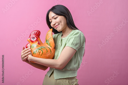 Asian woman in green top smiling warmly while hugging a large colorful ceramic rooster against a pink background
