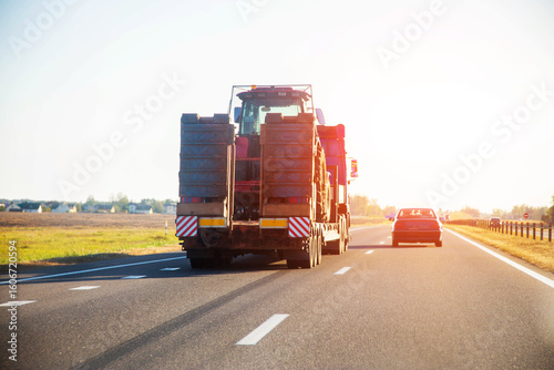 Fototapeta Naklejka Na Ścianę i Meble -  Transportation of a tractor by a truck with a lowboy semitrailer on a highway against the backdrop of a sunset in summer. Transportation of oversized special equipment, industry. Copy space for text