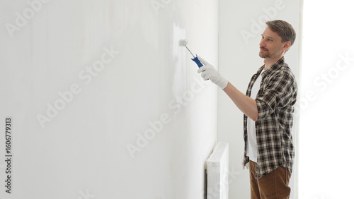 Middle aged man wearing checked shirt and protective gloves, is painting a white wall with a paint roller, working on a home renovation project, creating a fresh and clean look