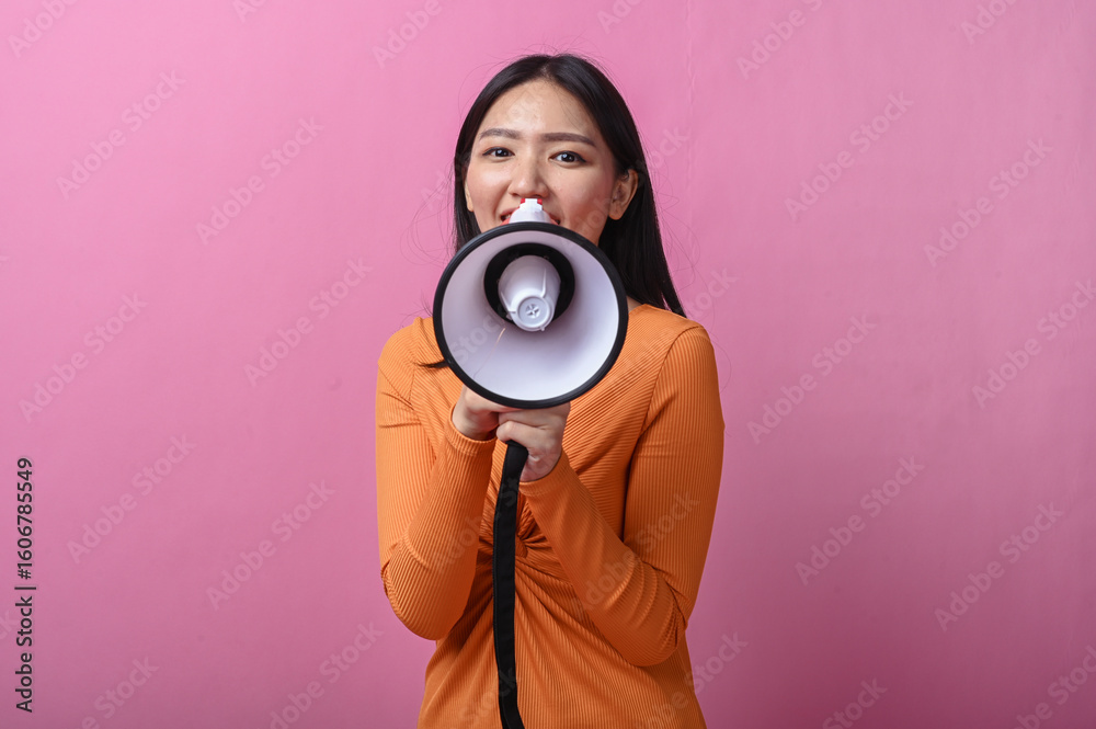 Fototapeta premium Asian woman with long black hair wearing an orange dress holding a megaphone directly facing the camera, standing against pink background, symbolizing voice, announcement, or call to action