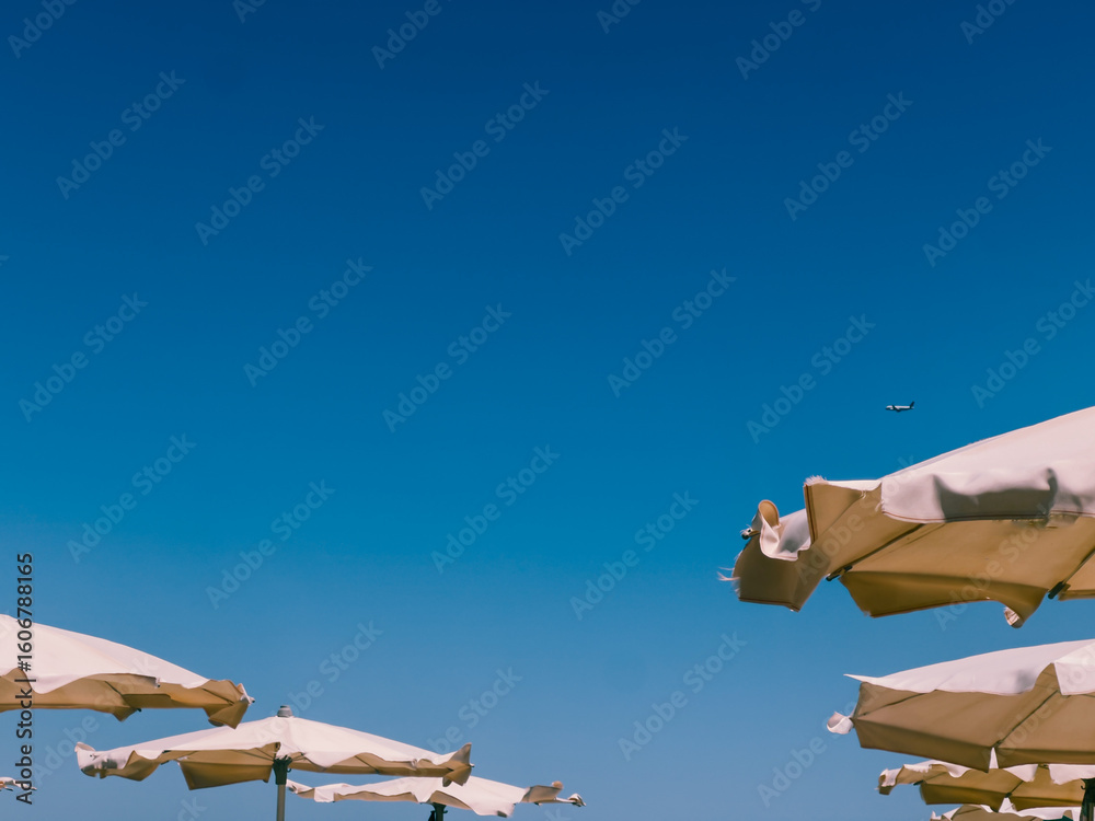 Fototapeta premium Tops of beach umbrellas with a plane flying in the distance, captured at Finikoudes Beach in Larnaca, Cyprus. Concept of summer vacation and seaside travel on the Mediterranean
