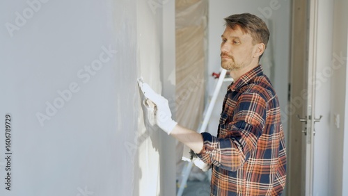 Male construction worker wearing protective gloves smoothing plaster onto wall surface during home renovation using professional trowel tool