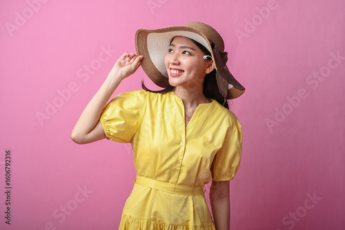 Asian woman in a yellow dress and wide-brim hat smiling softly while touching her hat, posing gracefully against a plain pink background