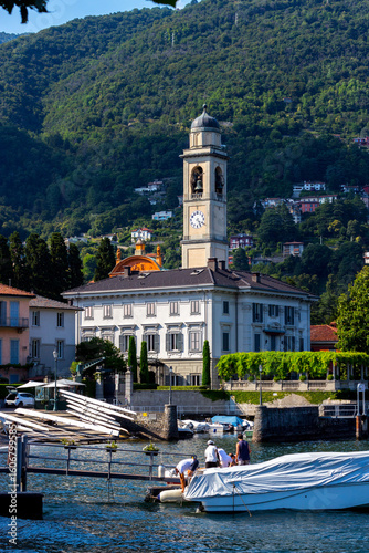 View of the town of Cernobbio on Lake Como, Lombardy. People on boats in the lake and a church with a clock in the background. Province of Como.